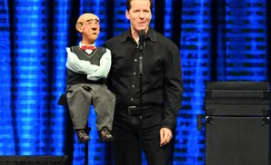 A ventriloquist and his puppet stand on stage with a blue backdrop.