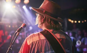 Musician in hat and plaid shirt performs on stage with microphone and blurred crowd.