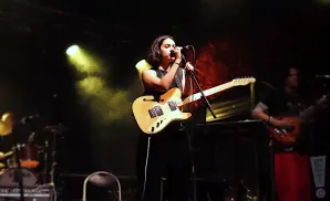 A woman sings and plays a light-colored electric guitar on stage, lit by spotlights.