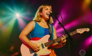 A woman with blonde hair plays a pink electric guitar on stage, illuminated by colorful lights.