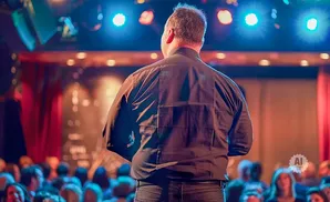 A man in a dark shirt and jeans stands on a stage, facing away from the camera, speaking to an audience.