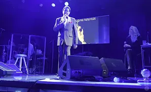 Wayne Newton on stage, holding a microphone, with a large screen displaying his album art behind him.