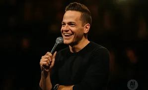 A man with a microphone, wearing a black shirt, smiles widely on a dark stage.