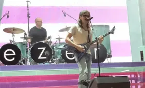 Musician with mullet and glasses plays electric guitar onstage with band, drums visible.