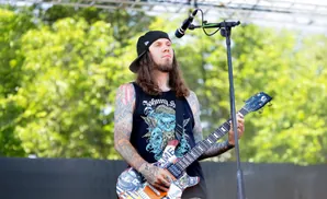 A guitarist with tattoos and a black cap plays a decorated guitar on stage.