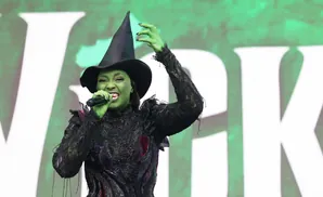 A woman in green face paint, a black witch hat, and a dark, ornate costume sings into a microphone.