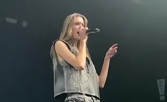 A woman sings into a microphone on stage, wearing a gray vest and patterned pants.