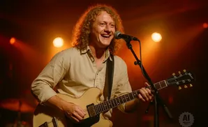 A smiling man with curly blonde hair plays a tan electric guitar and sings into a microphone on a stage with orange lights.