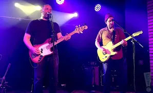 A band performs on a dimly lit stage with purple spotlights. Two guitarists are visible.