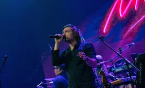 A male singer in a black shirt holds a microphone on stage, with pink and blue lights in the background.
