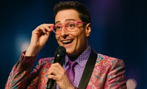 A man with pink glasses and a colorful jacket holds a microphone.