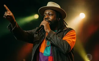 A man in a hat and jacket sings into a microphone, gesturing with his hand.