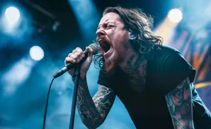 A tattooed man with long hair sings passionately into a microphone on a dimly lit stage.