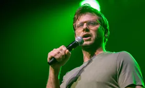 A man in glasses and a green t-shirt speaks into a microphone under green stage lighting.
