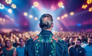 Man with embroidered jacket faces a blurred, cheering crowd under colorful stage lights.