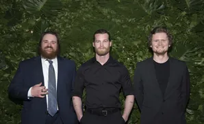 Three men in suits stand smiling in front of a green, leafy backdrop.