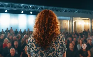 A woman with curly red hair faces away from the camera, speaking to a seated audience in a dimly lit venue.