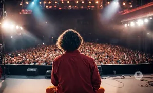 A person with curly hair in a red shirt sits on a stage facing a large, cheering crowd in a concert hall.