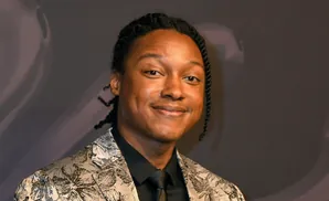 A Black man with braided hair wears a patterned suit and a black shirt and tie, smiling at the camera.