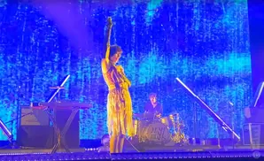 A singer in a fringe dress performs on a stage with a drummer behind her, set against a blue, textured backdrop.