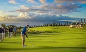 Golfer on a green prepares to putt, with teammates watching and a mansion on a distant hill.