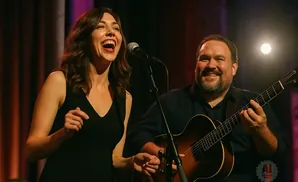 A female singer laughs into a microphone as a man plays guitar beside her.