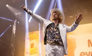 A male singer in a white cowboy hat and suit performs on stage with lights and a banner behind him.