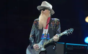 A male guitarist with a long blonde beard, wearing a hat and sunglasses, plays a decorated guitar on stage.