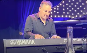 A man plays a Yamaha keyboard in front of a grand piano, with a blurred bottle in the foreground.