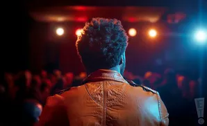 Back view of a person with curly hair wearing a leather jacket on a stage with red and blue lights.