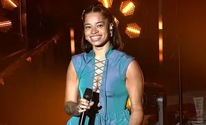 Singer Ella Mai smiles while performing on stage in a blue lace-up top and gold earrings.