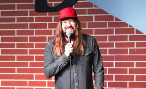 Comedian with long brown hair, glasses, and a red baseball cap holds a microphone in front of a brick wall.