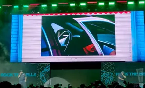 A person on stage performs in front of a large screen displaying a man inside a car.