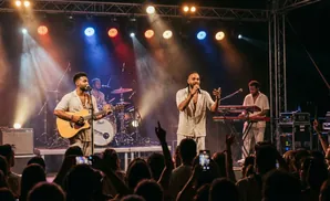A band performs on stage with bright lights. A singer holds a microphone and gestures, while a guitarist plays an acoustic guitar.
