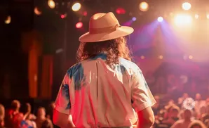 Man with long hair wearing a hat and a tie-dye shirt on stage under bright lights.