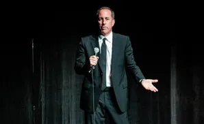 Jerry Seinfeld stands on stage in a black suit, holding a microphone and gesturing with his hand.