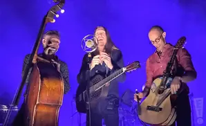 Three musicians perform on a stage with a blue background. The musician on the left plays a double bass and harmonica. The middle musician sings into a …