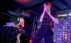 A male singer holds a microphone while a female keyboardist plays a keyboard on stage.