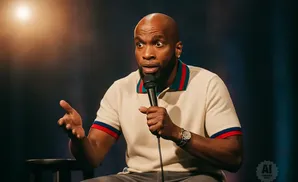 A Black man with a bald head speaks into a microphone while sitting on a stool.