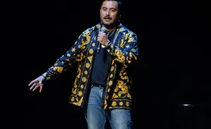 Comedian on stage wearing a black and gold patterned shirt and jeans, holding a microphone.