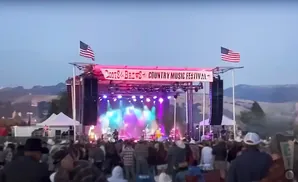 A country music festival stage with American flags and a large crowd gathered.