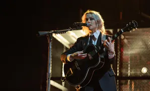 A woman with blonde hair plays a black guitar and sings into a microphone on stage.
