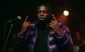 A man with braids in a purple puffer jacket and black shirt gestures with rock-on signs in front of a microphone.