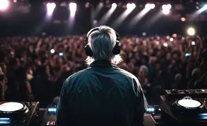 DJ with headphones plays for a large, blurred crowd at a concert, illuminated by bright stage lights.