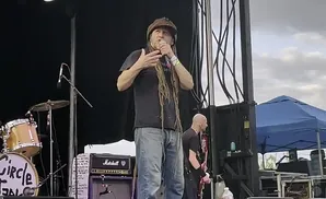 A dreadlocked man in a hat speaks into a microphone on a stage.