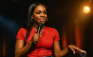 A Black woman in a red shirt holds a microphone and speaks on stage under a spotlight.
