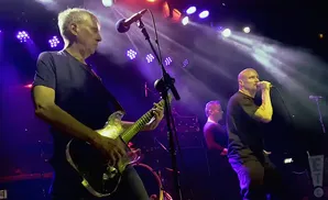 A bald man sings into a microphone on a stage, with a guitarist and another man behind him.