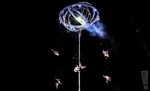Aerial performers hang from a luminous, spherical apparatus against a black sky.