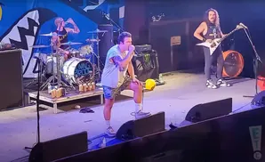 A band performs on stage, with a singer in the foreground and a guitarist and drummer behind him.