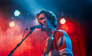 A male singer performs on stage, bathed in red and blue lights, with a microphone in front of him.